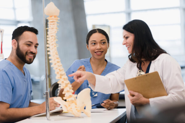 Students learn about human spine from female medical school professor. Two young adult interns learn about the human spine from their female mid adult medical school teacher.